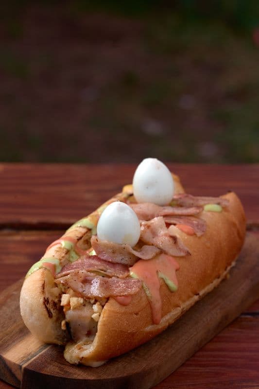 Loaded hot dog with bacon, two quail eggs, and colorful sauces on a wooden board.