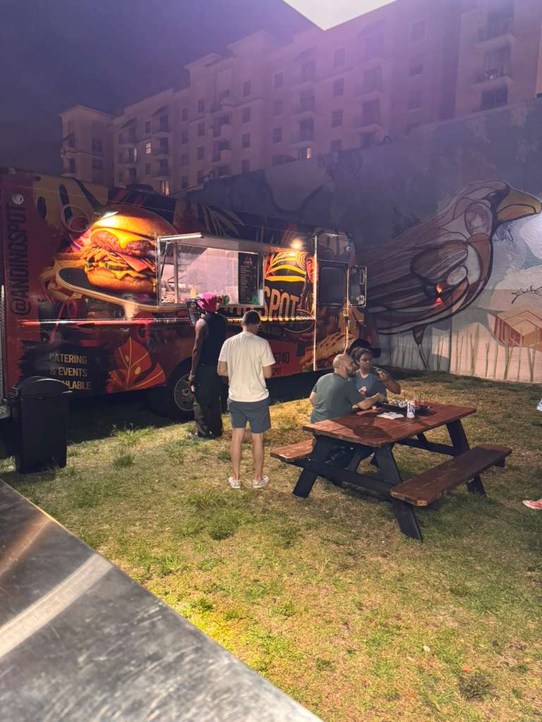 People order from a vibrant food truck at night beside a wooden picnic table.