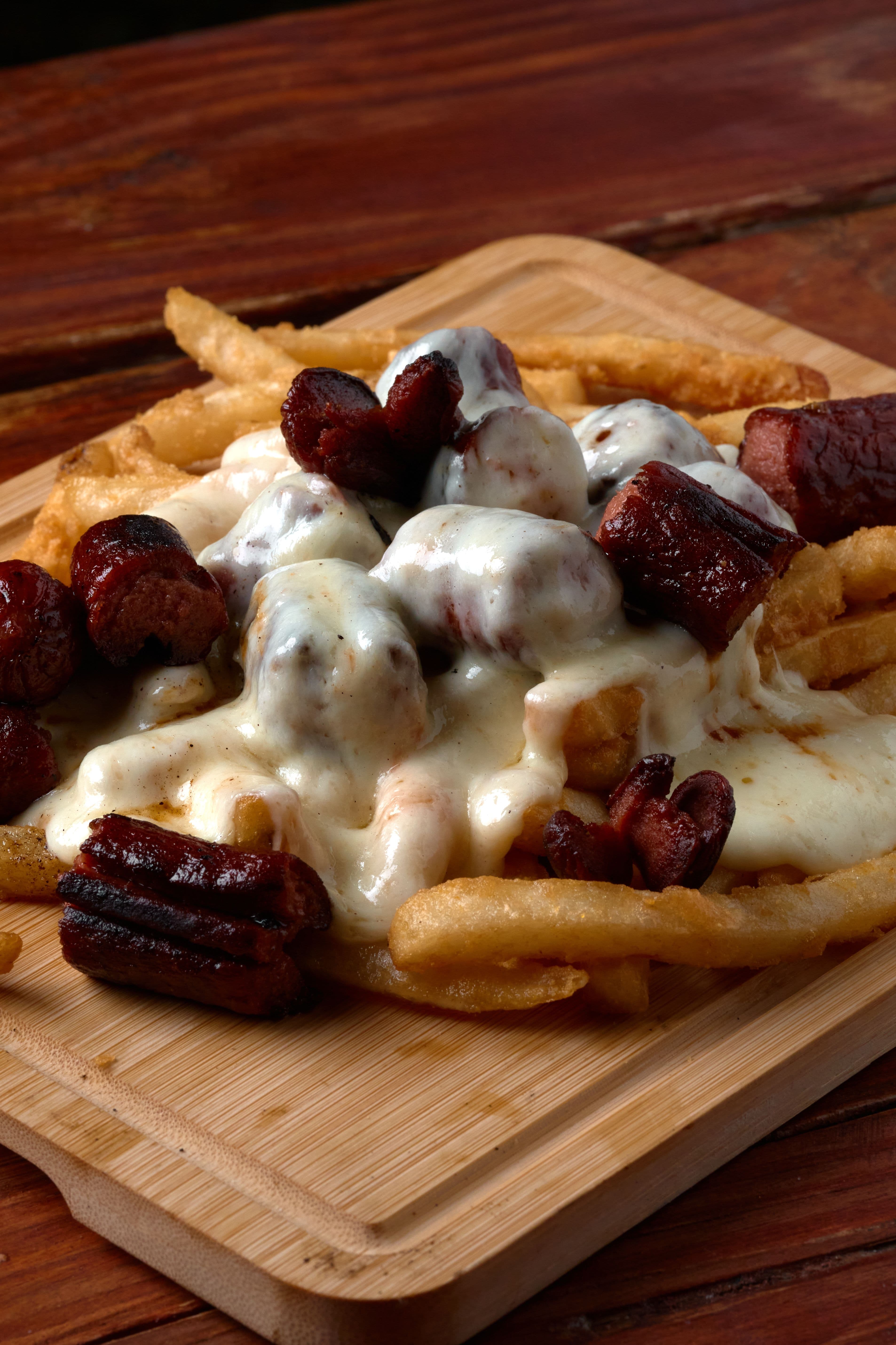 French fries topped with melted white cheese and grilled sausage on a wooden board.