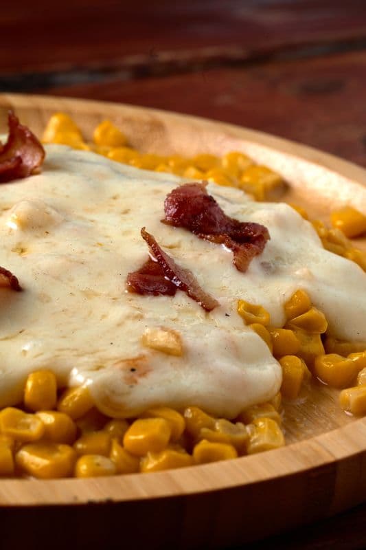 Melted white cheese and crispy bacon bits over yellow corn kernels on a wooden dish.